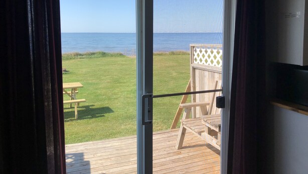 A view from a room with a sliding glass door to a deck overlooking a grassy area with a wooden bench and a wooden structure.