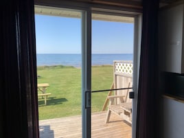 A view from a room with a sliding glass door to a deck overlooking a grassy area with a wooden bench and a wooden structure.