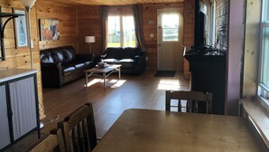 A cozy cabin interior with a wooden floor, a leather sofa, a dining table, and a kitchen area with a stove.