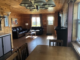 A cozy cabin interior with a wooden floor, a leather sofa, a dining table, and a kitchen area with a stove.