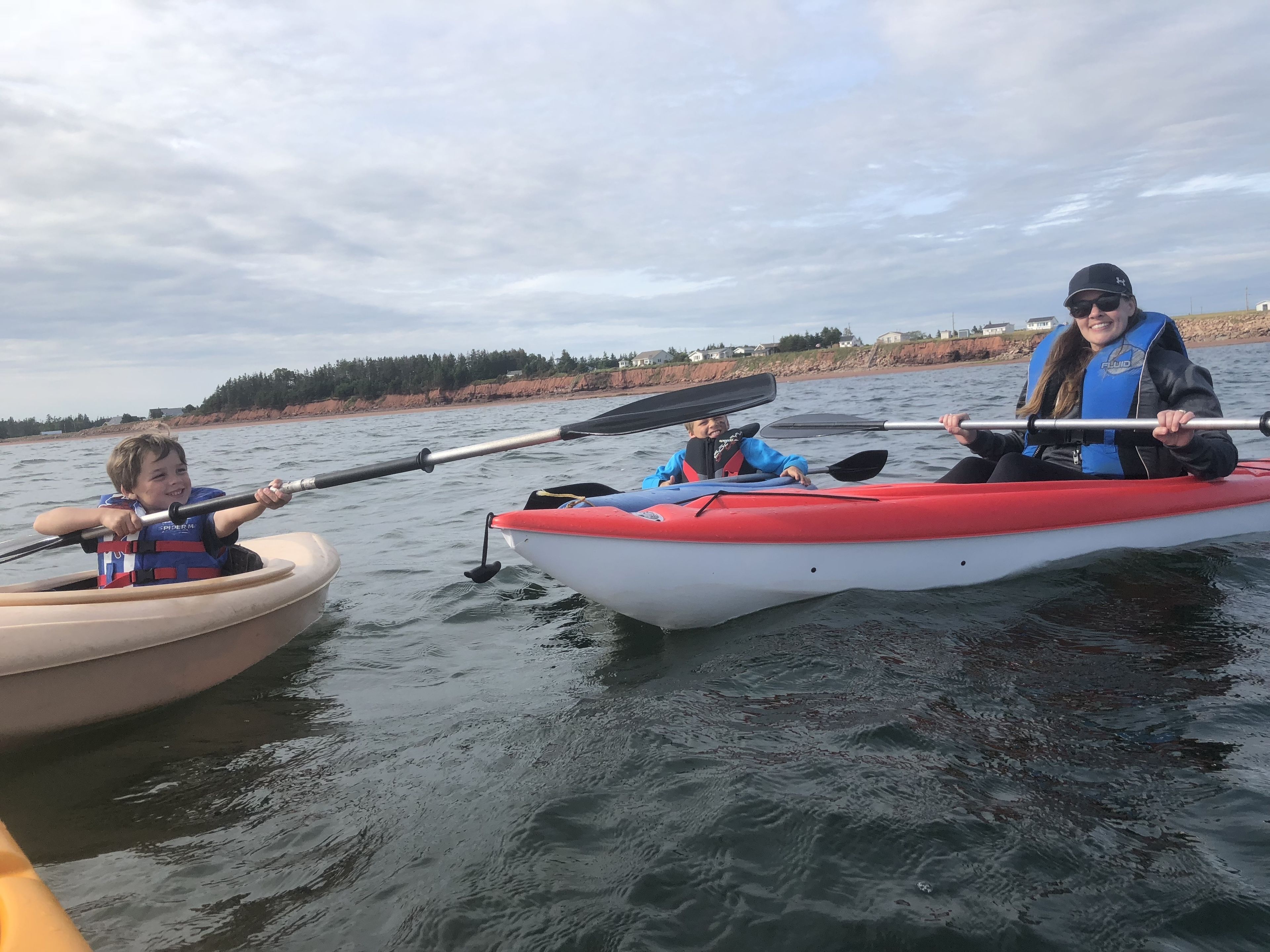 Three people kayaking on a calm body of water with a shoreline in the background.