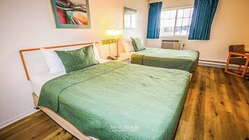A hotel room with two beds, a wooden floor, a window with curtains, a painting on the wall, and a lamp.