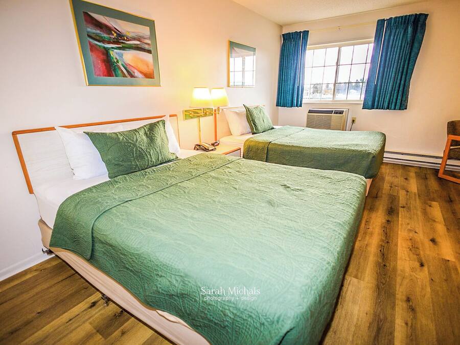 A hotel room with two beds, a wooden floor, a window with curtains, a painting on the wall, and a lamp.