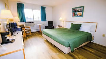 A hotel room with a bed, a desk with a lamp, a chair, a small table, and a window with curtains.