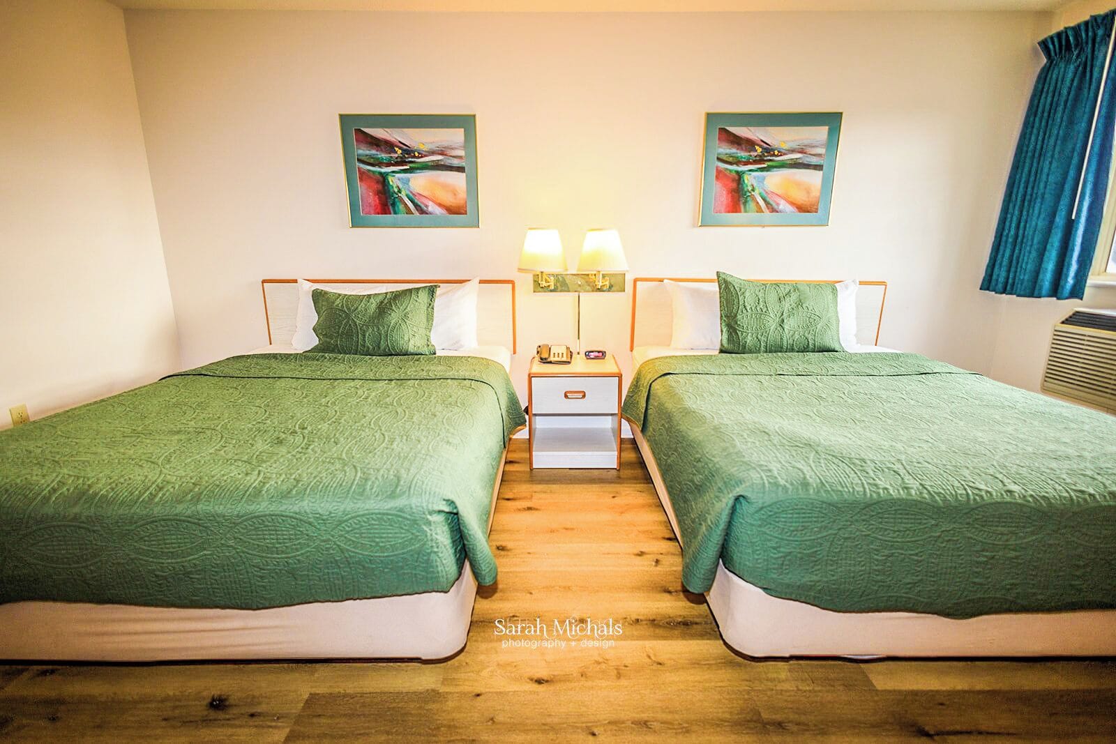 Ein Hotelzimmer mit zwei Betten, jeweils mit grüner Bettwäsche, einem Nachttisch mit Lampe sowie zwei gerahmten abstrakten Bildern an der Wand.