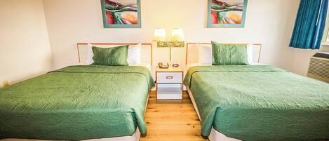Ein Hotelzimmer mit zwei Betten, jeweils mit grüner Bettwäsche, einem Nachttisch mit Lampe sowie zwei gerahmten abstrakten Bildern an der Wand.