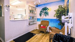 Une pièce comprenant un comptoir d’accueil, des chaises en osier, une petite table, une fresque murale représentant une scène de plage et une étagère contenant divers objets.