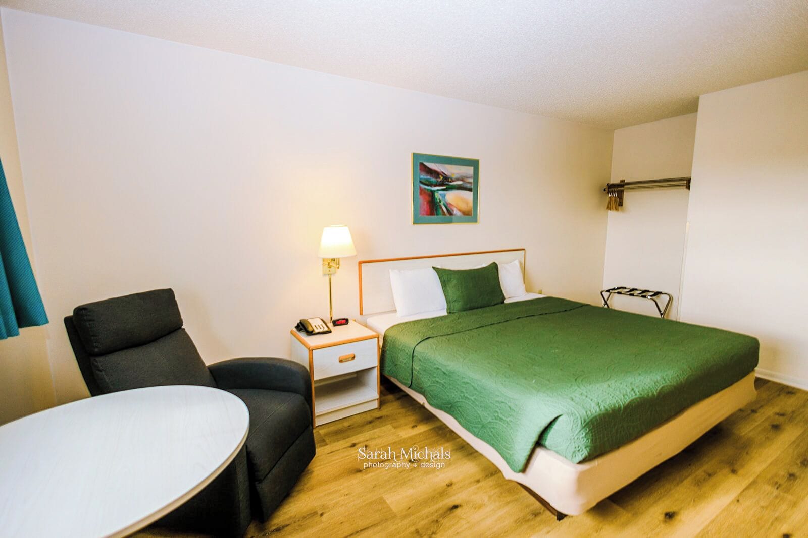 A hotel room with a bed, a chair, a bedside table with a lamp, and a small table.