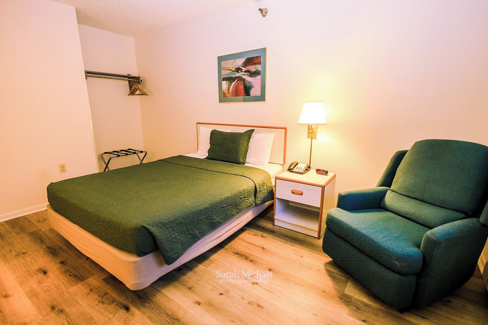 A room with a bed, a nightstand with a lamp, a chair, and a painting on the wall.