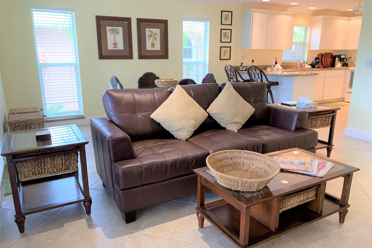 A living room with a brown leather sofa, two side tables, a coffee table, and a kitchen in the background.
