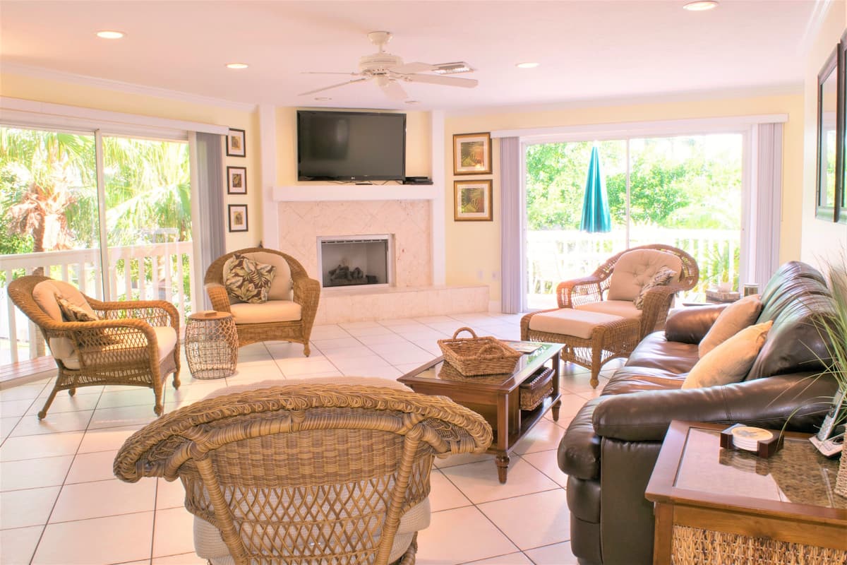A spacious living room with wicker furniture, a leather sofa, a flat-screen TV, and a fireplace.