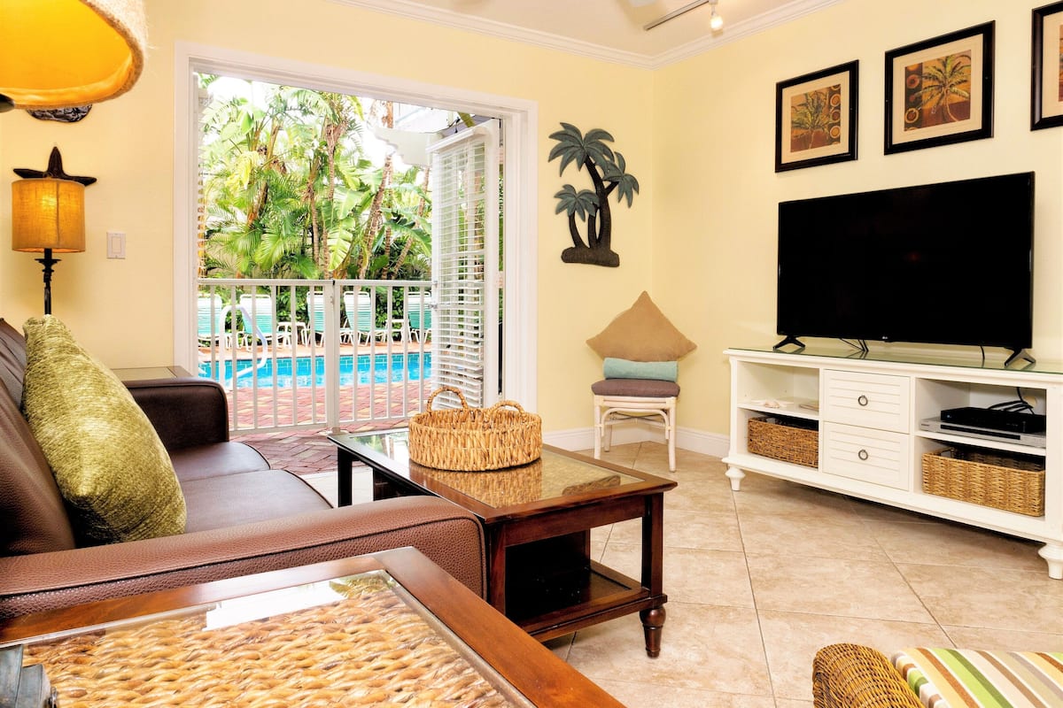 A living room with a sofa, a television, a coffee table, and a view of a pool.