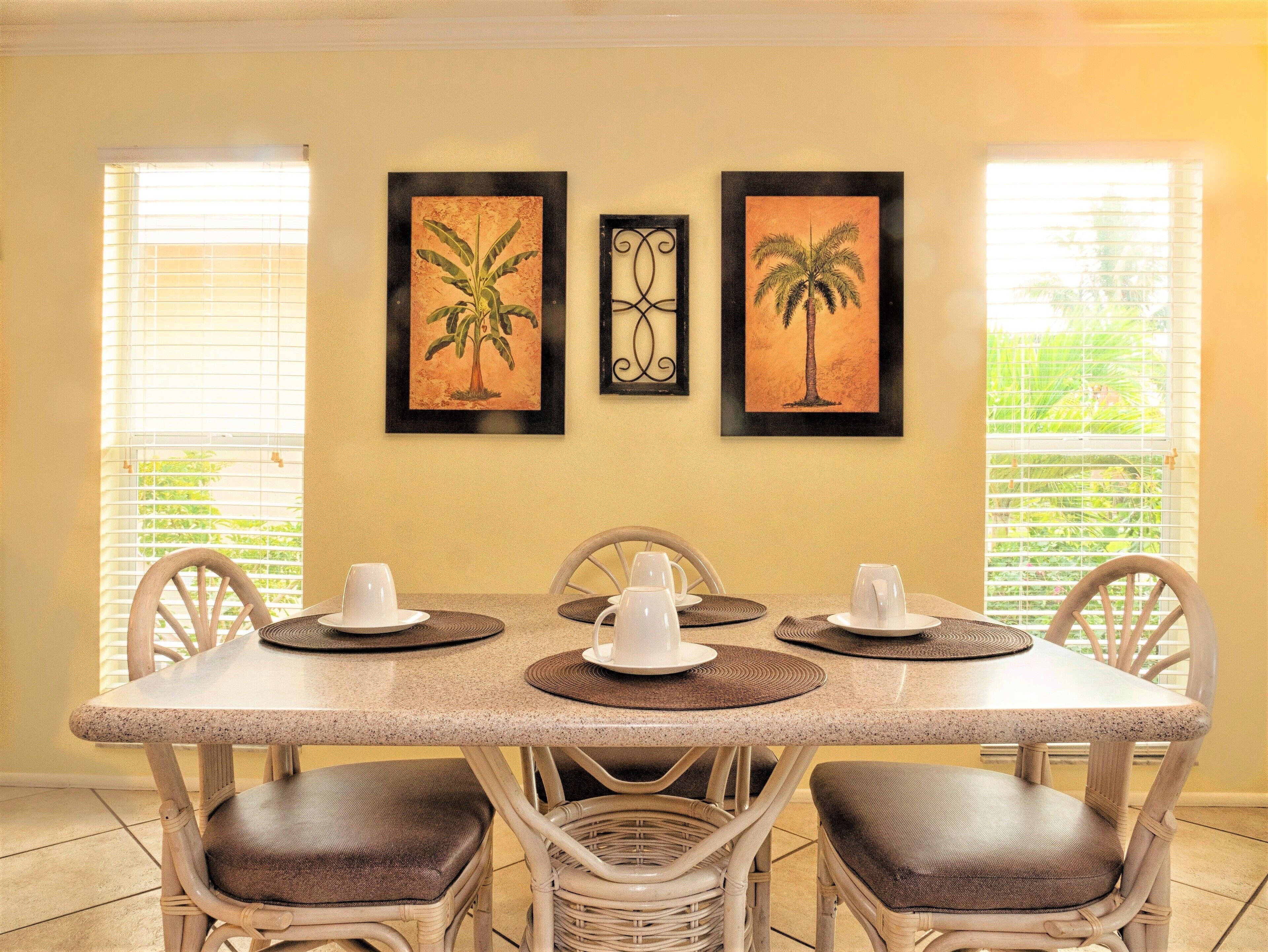 A dining area with a table set for four, framed artwork on the wall, and large windows with blinds.