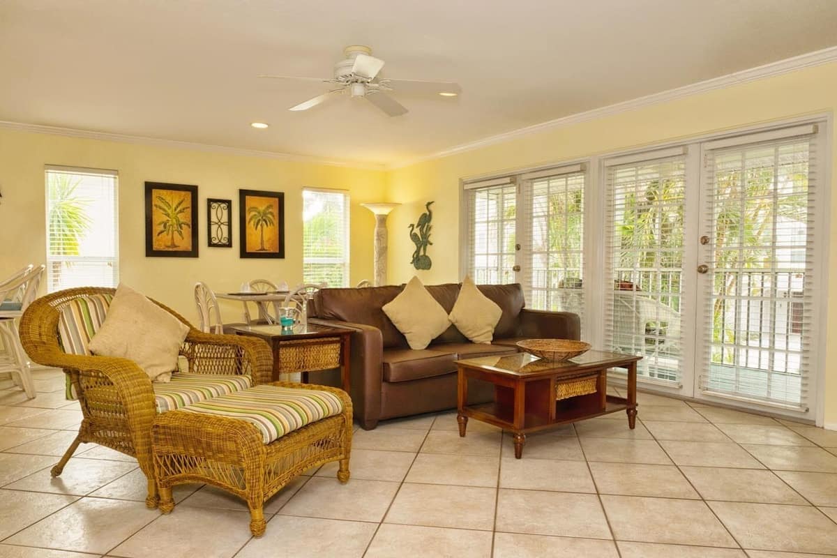 A living room with a wicker sofa, a brown leather couch, and a wooden coffee table. There are framed pictures on the wall, a ceiling fan, and a window with blinds.