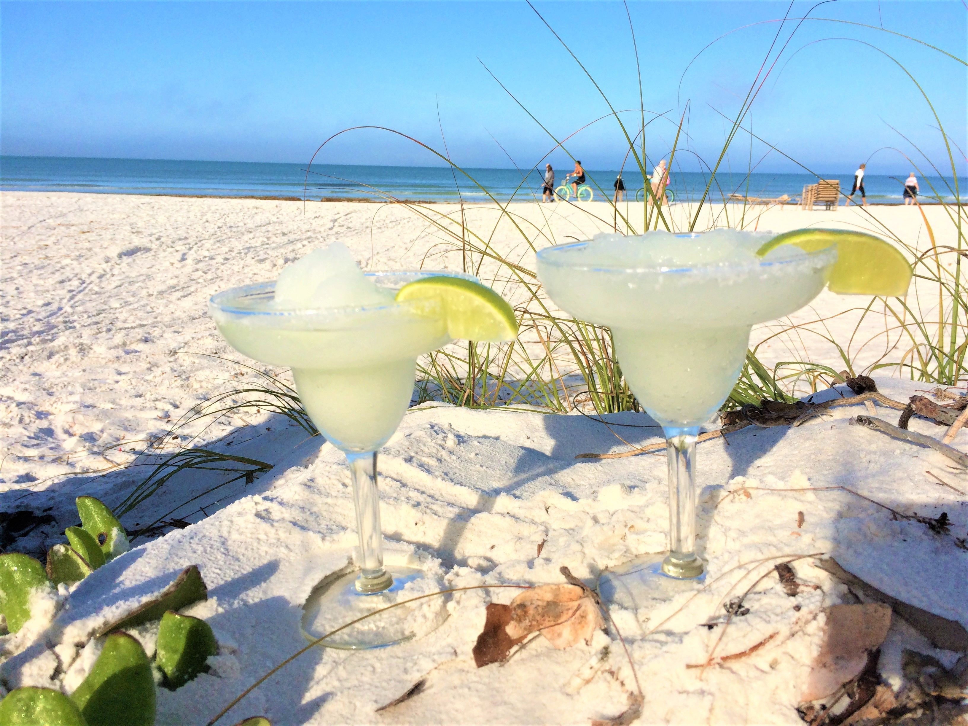 Two margaritas on a sandy beach with a clear view of the ocean and beachgoers in the background.