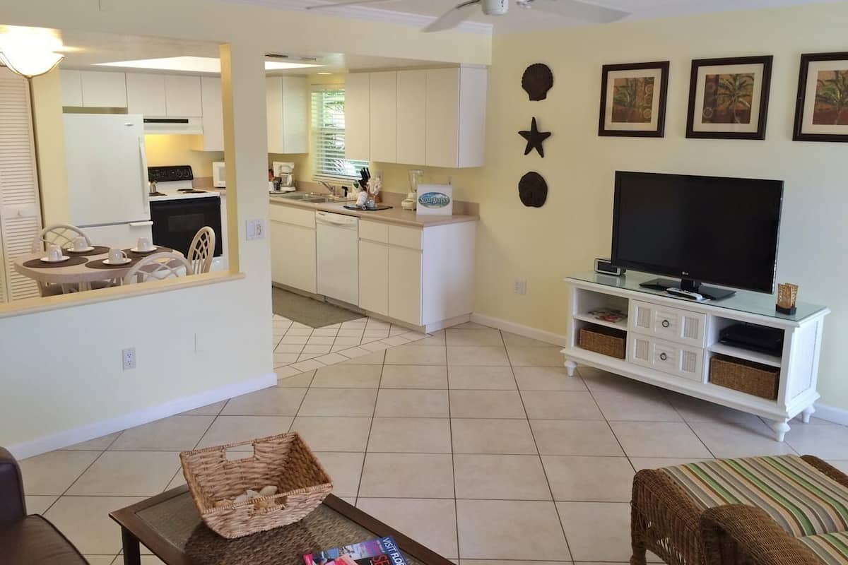 A living room with a TV, a dining area with a table and chairs, a kitchen with white cabinets, and a ceiling fan.