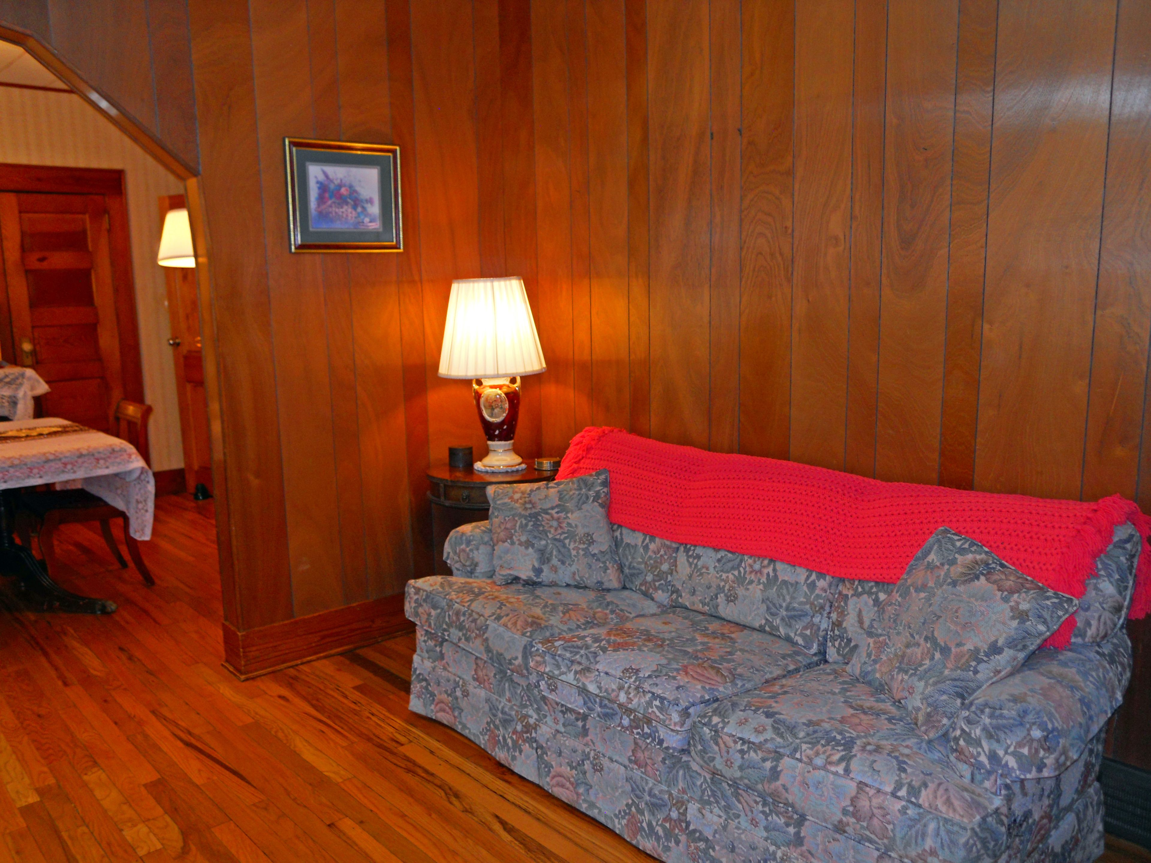 A room with wooden paneling, a floral sofa, a red blanket, a table with a lamp, and a framed picture on the wall.