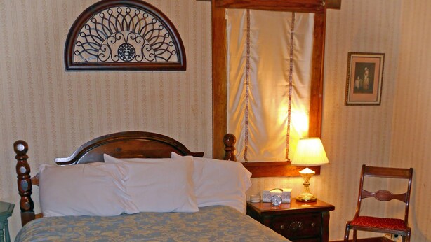 A neatly made bed with a patterned bedspread, a wooden headboard, a bedside table with a lamp, a chair, and a wall-mounted decorative piece. - Little River Inn Motel & Guest House (Leon)