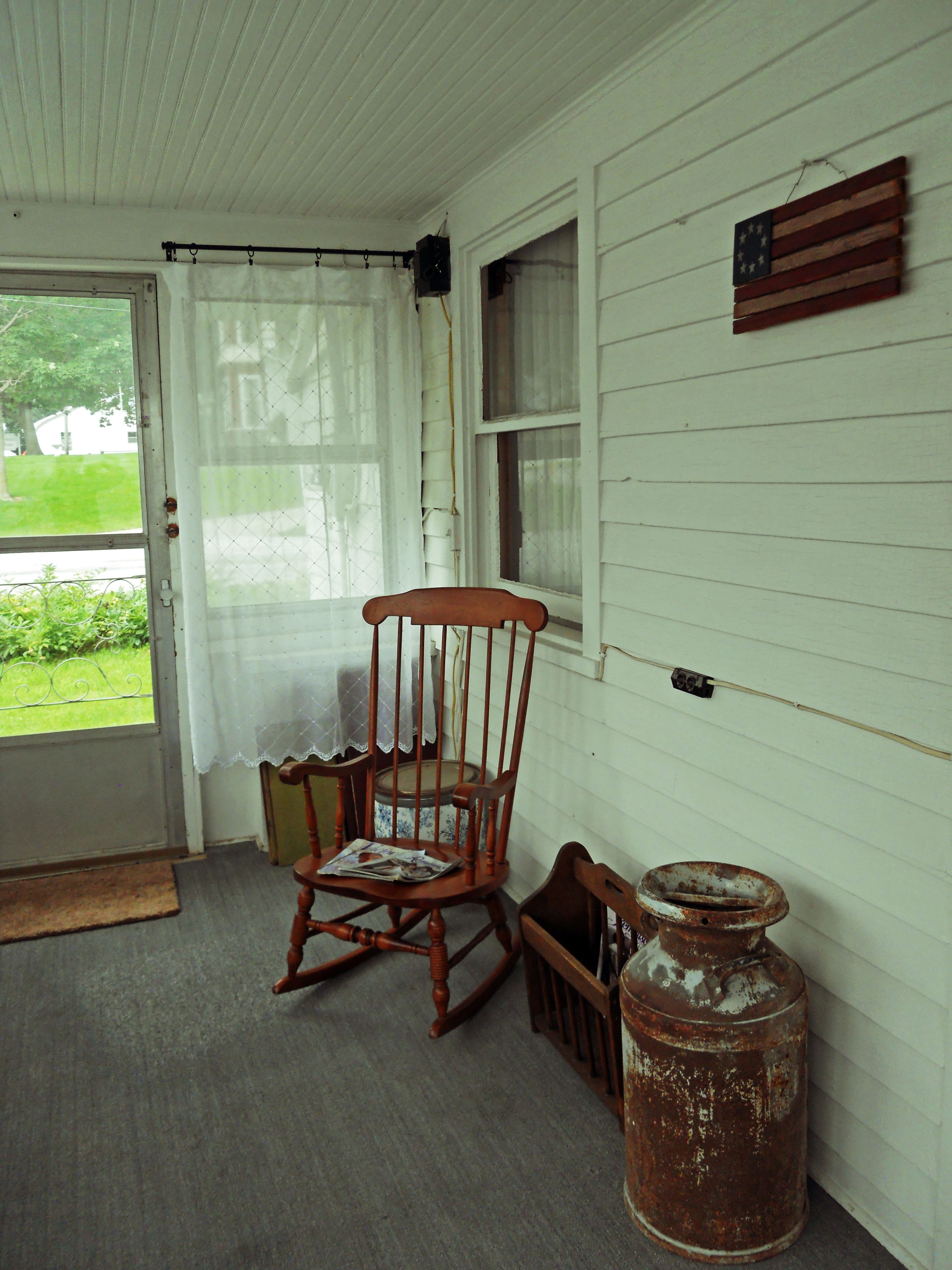 A porch with a rocking chair, a large metal container, and a window with a curtain.