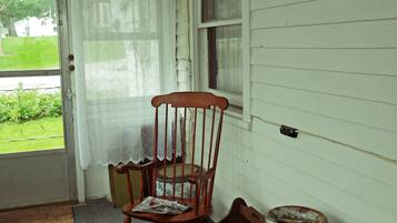 A porch with a rocking chair, a large metal container, and a window with a curtain.