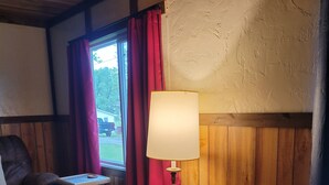 A cozy room with a leather armchair, a small wooden table with a lamp, and a view of the outdoors through a window with red curtains. - Little River Inn Motel & Guest House (Leon)