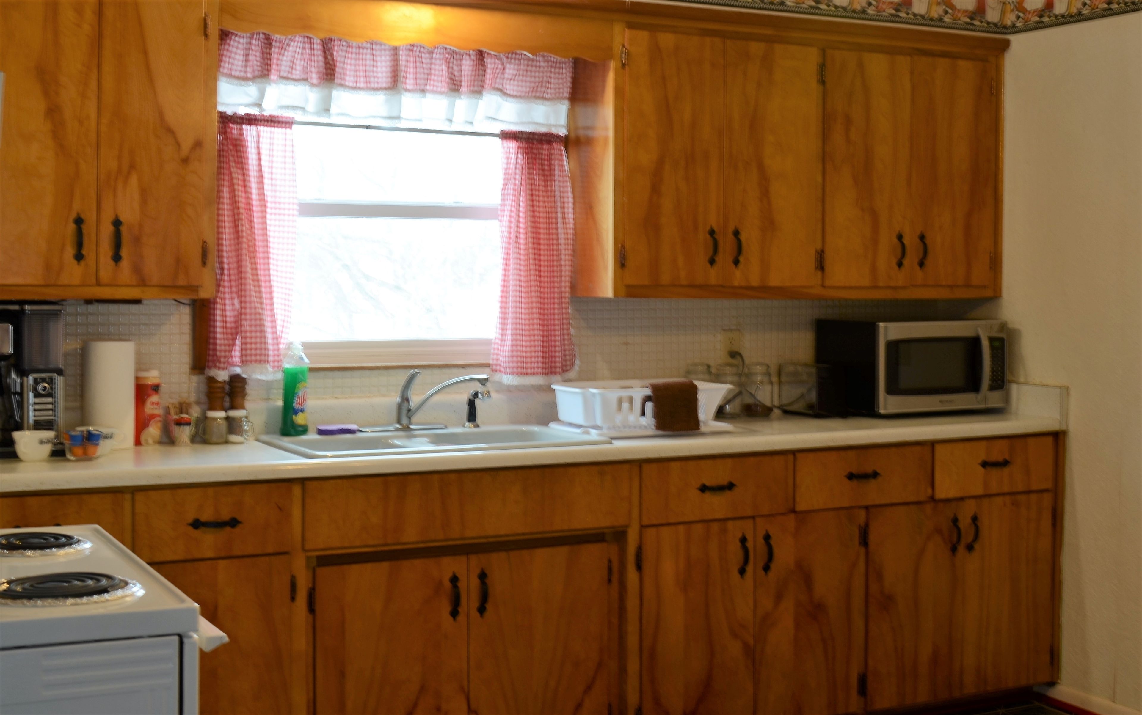 A kitchen with wooden cabinets, a white stove, a microwave, and a window with a checkered curtain.
