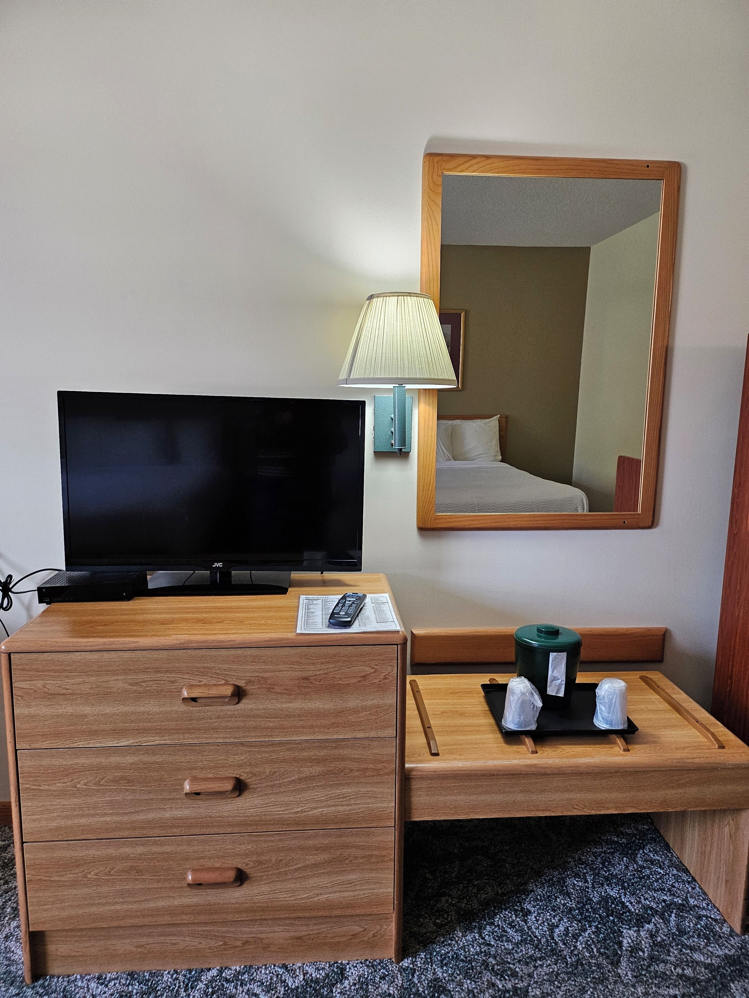Habitación de hotel con un escritorio de madera, televisor, lámpara, espejo y una pequeña mesa con dos tazas y un recipiente verde.