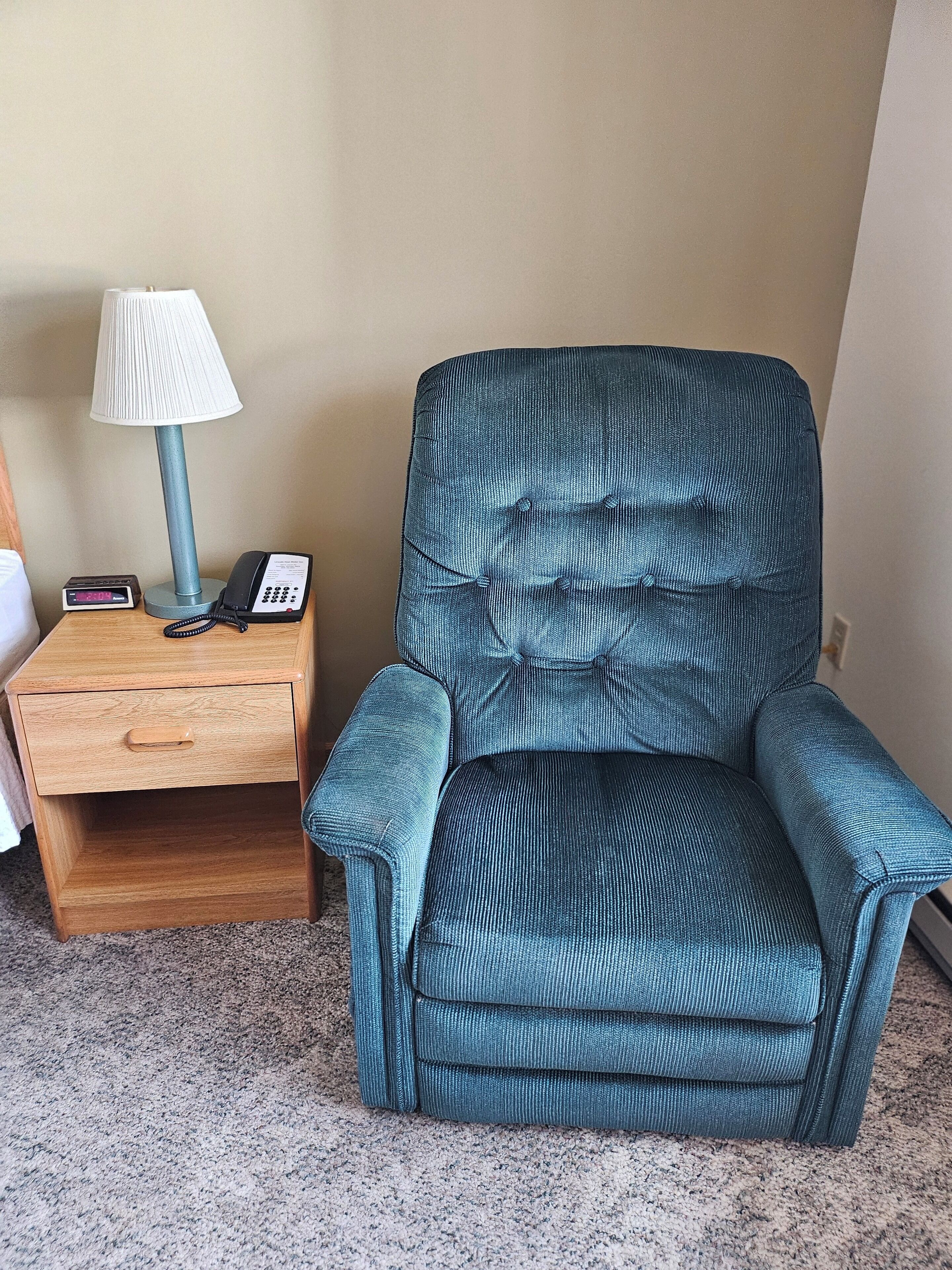 Ein Zimmer mit einem Holznachttisch, einer Lampe, einem Telefon und einem blauen Polsterarmlehnstuhl.