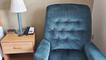 Ein Zimmer mit einem Holznachttisch, einer Lampe, einem Telefon und einem blauen Polsterarmlehnstuhl.