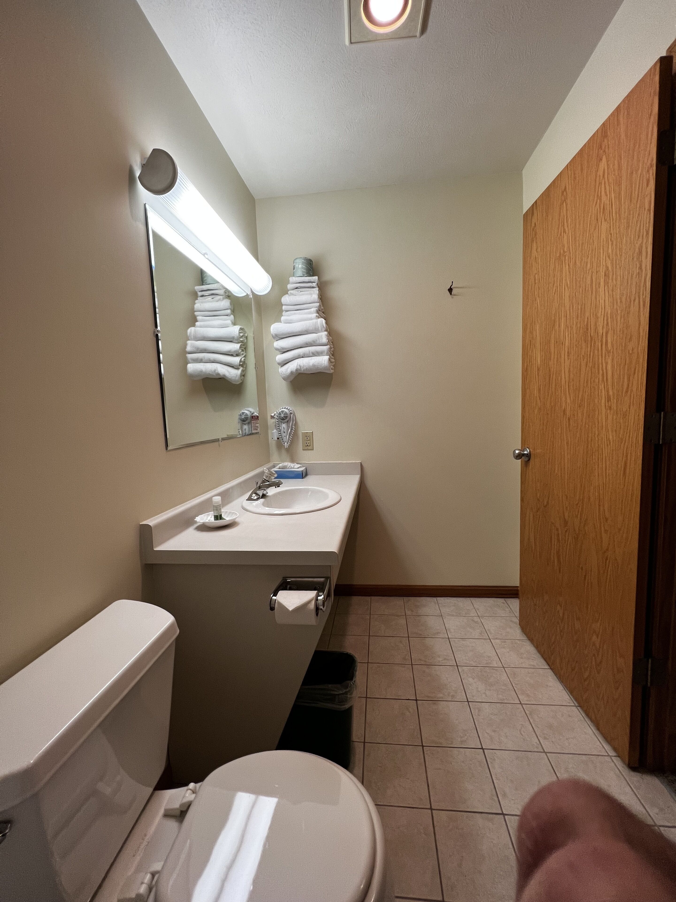 Baño con inodoro, lavamanos y espejo. Hay toallas colgadas en la pared y un cesto de basura.