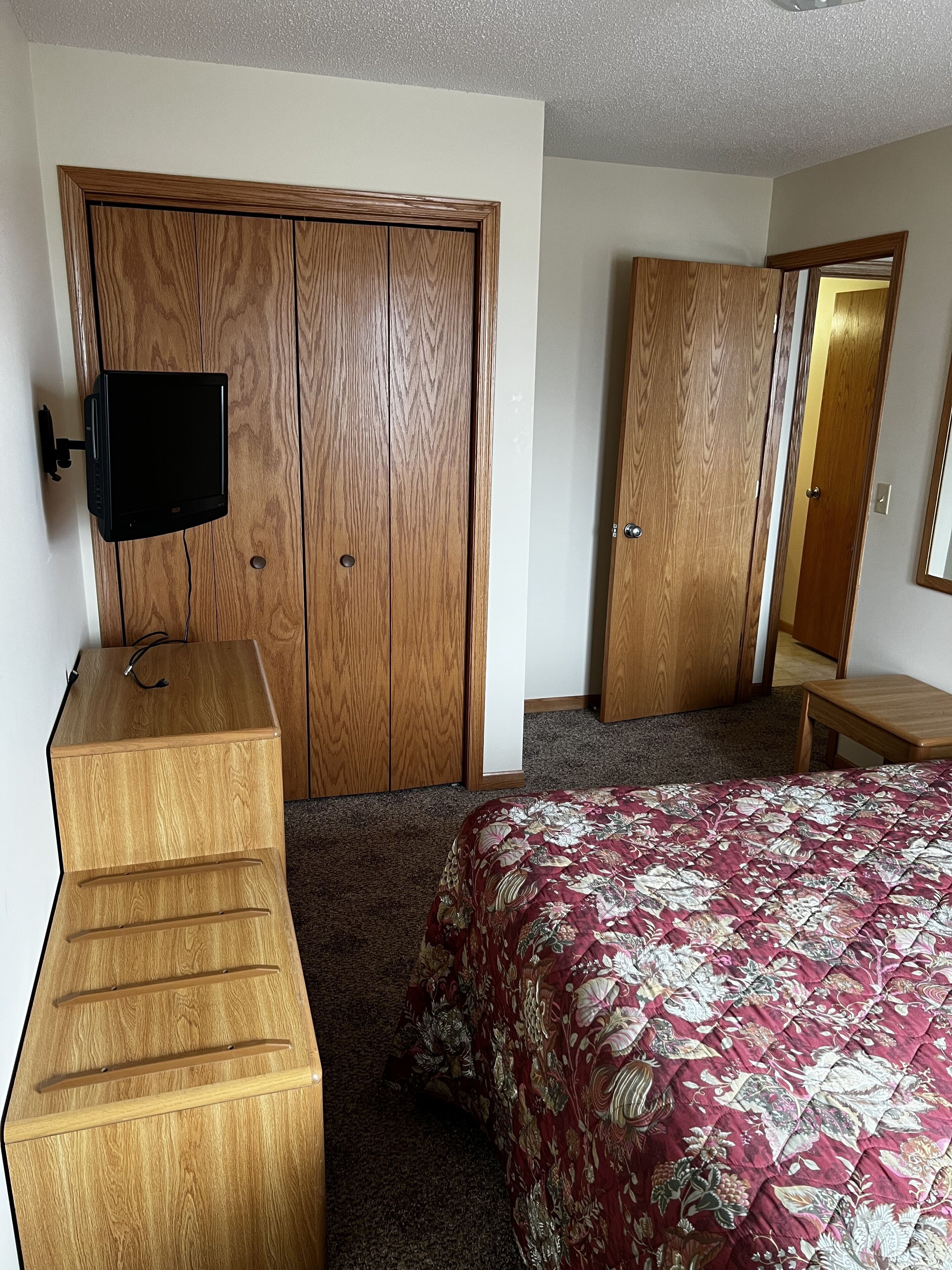 A bedroom with a wooden dresser, a bed with a floral bedspread, a television on the wall, and a wooden wardrobe.
