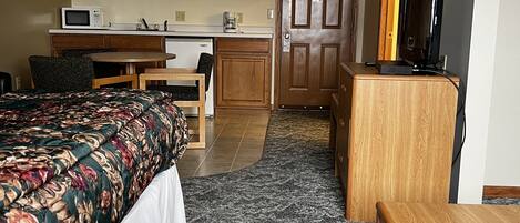 A compact hotel room with a bed, a desk, a kitchenette, and a television.