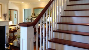 Una imponente escalera con balaustres blancos y peldaños de madera oscura, que conduce a un descansillo con chimenea y un arreglo floral.