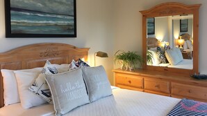 Una cama bien hecha con almohadones decorativos, un cuadro grande de un paisaje marino y un espejo que refleja el interior de la habitación.
