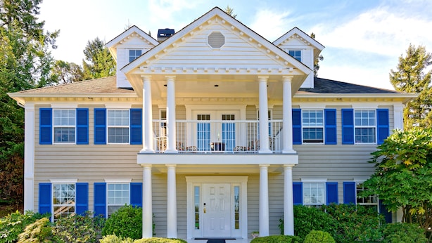 Una casa de dos pisos con un balcón central, columnas blancas y contraventanas azules.