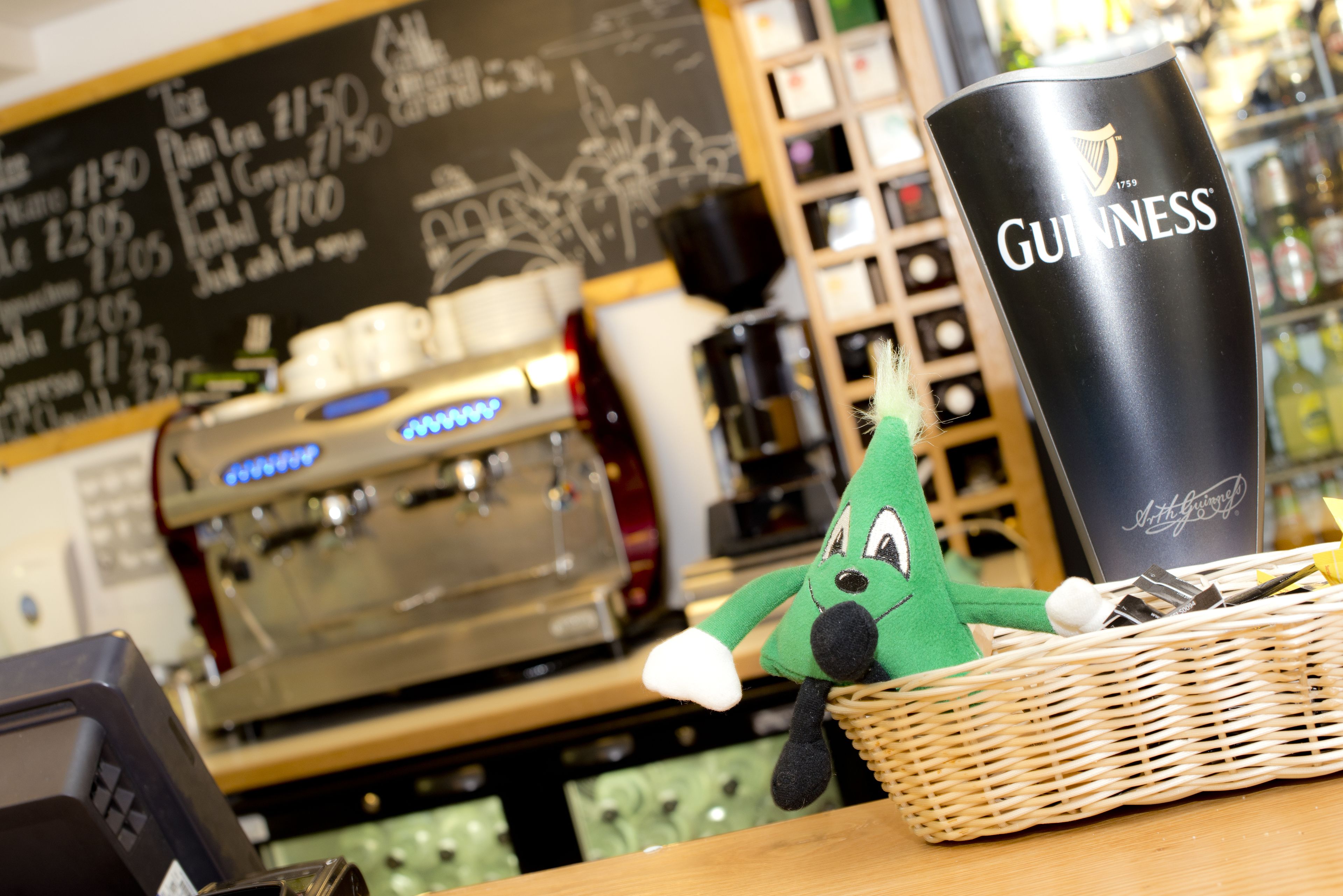 A green plush toy in a wicker basket, a black Guinness glass, and a coffee machine in a cafe setting.