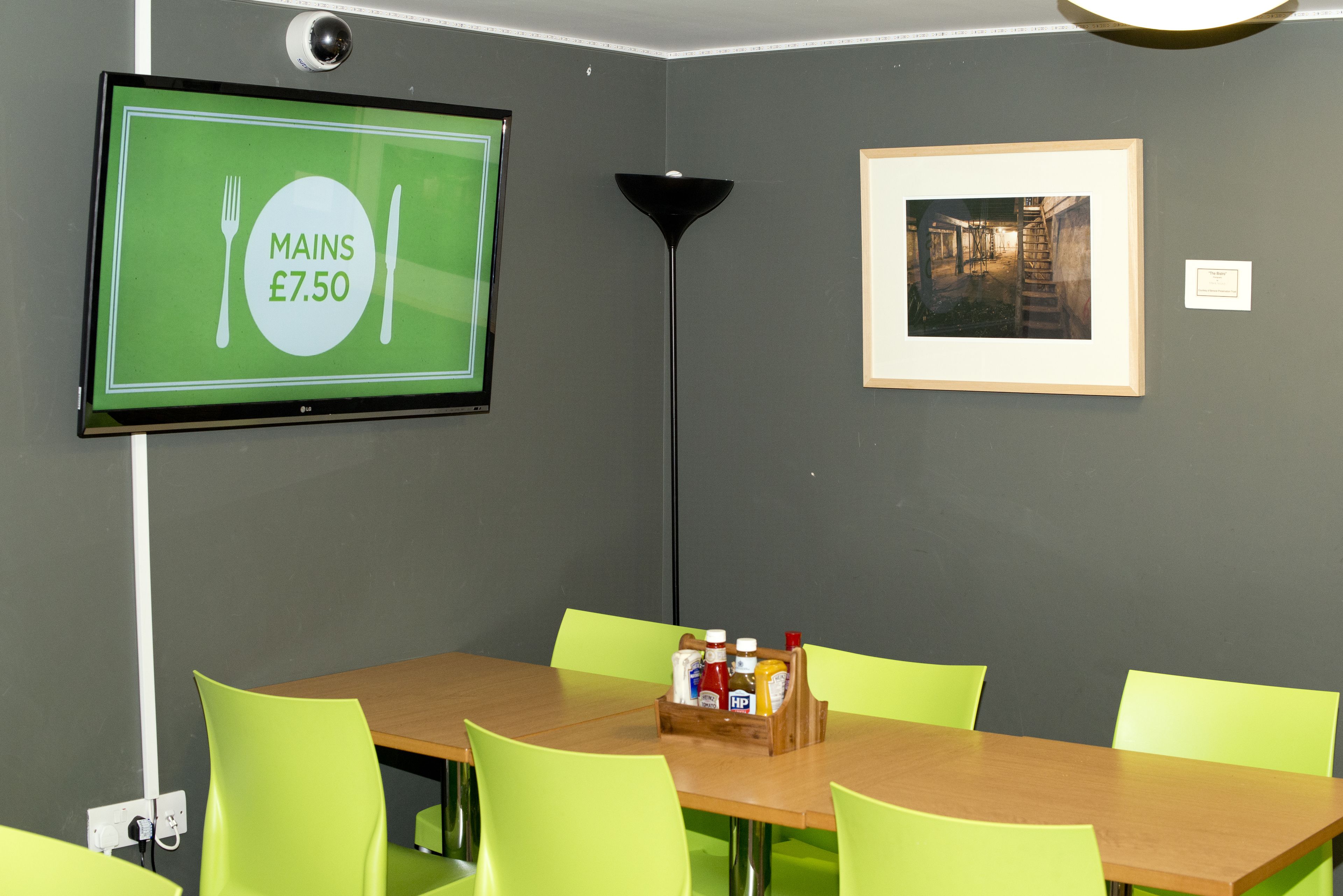 Un área de comedor con una mesa, sillas, un televisor mostrando el precio de una comida y un cuadro en la pared.