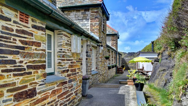 A narrow stone pathway flanked by buildings with stone walls and traditional windows. There are outdoor seating areas with tables and chairs, and potted plants along the path. - YHA Boscastle - Hostel (Boscastle)