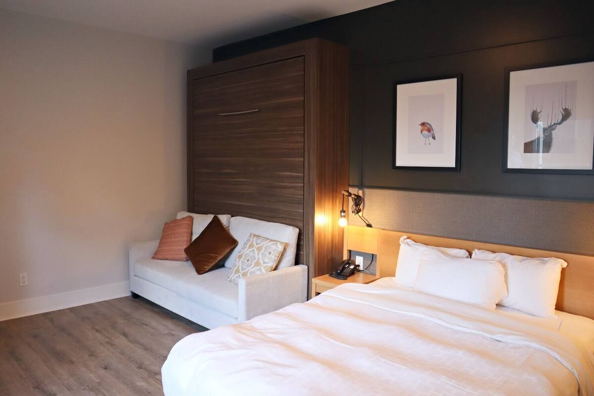 A hotel room with a bed, a sofa, a nightstand, and a wall-mounted lamp.