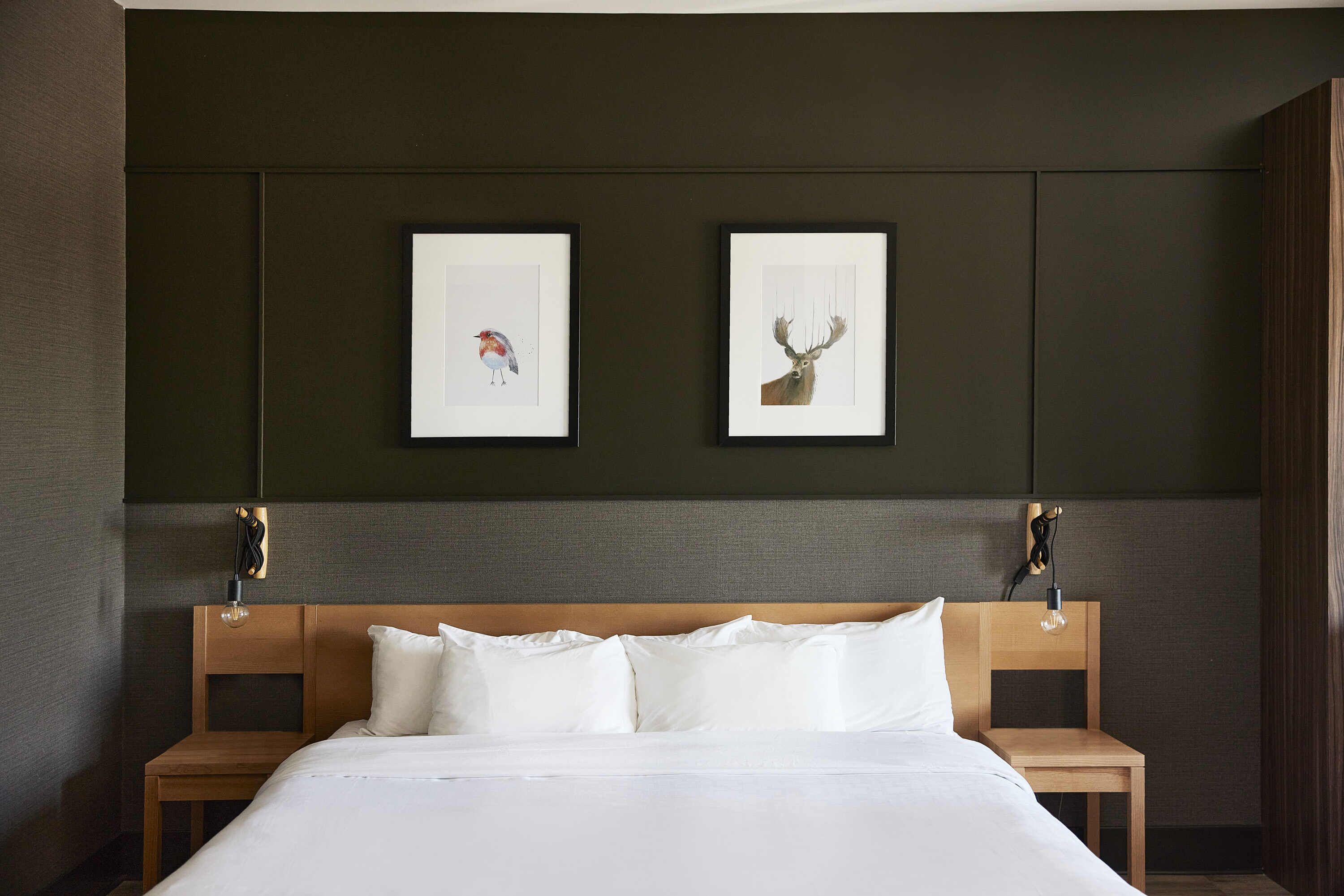 A neatly made bed with white linens, two framed pictures on the wall, and a wooden nightstand beside it.