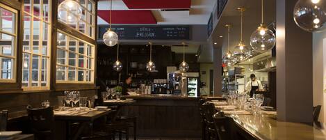 A restaurant interior with wooden tables and chairs, hanging pendant lights, and a bar area.