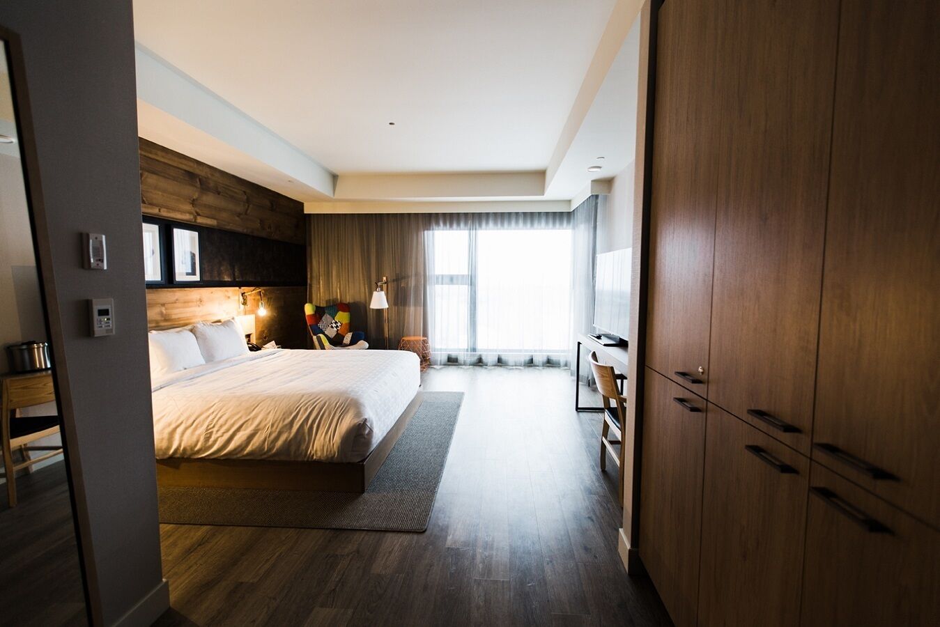 A modern hotel room with a large bed, wooden flooring, a desk, and a wardrobe.