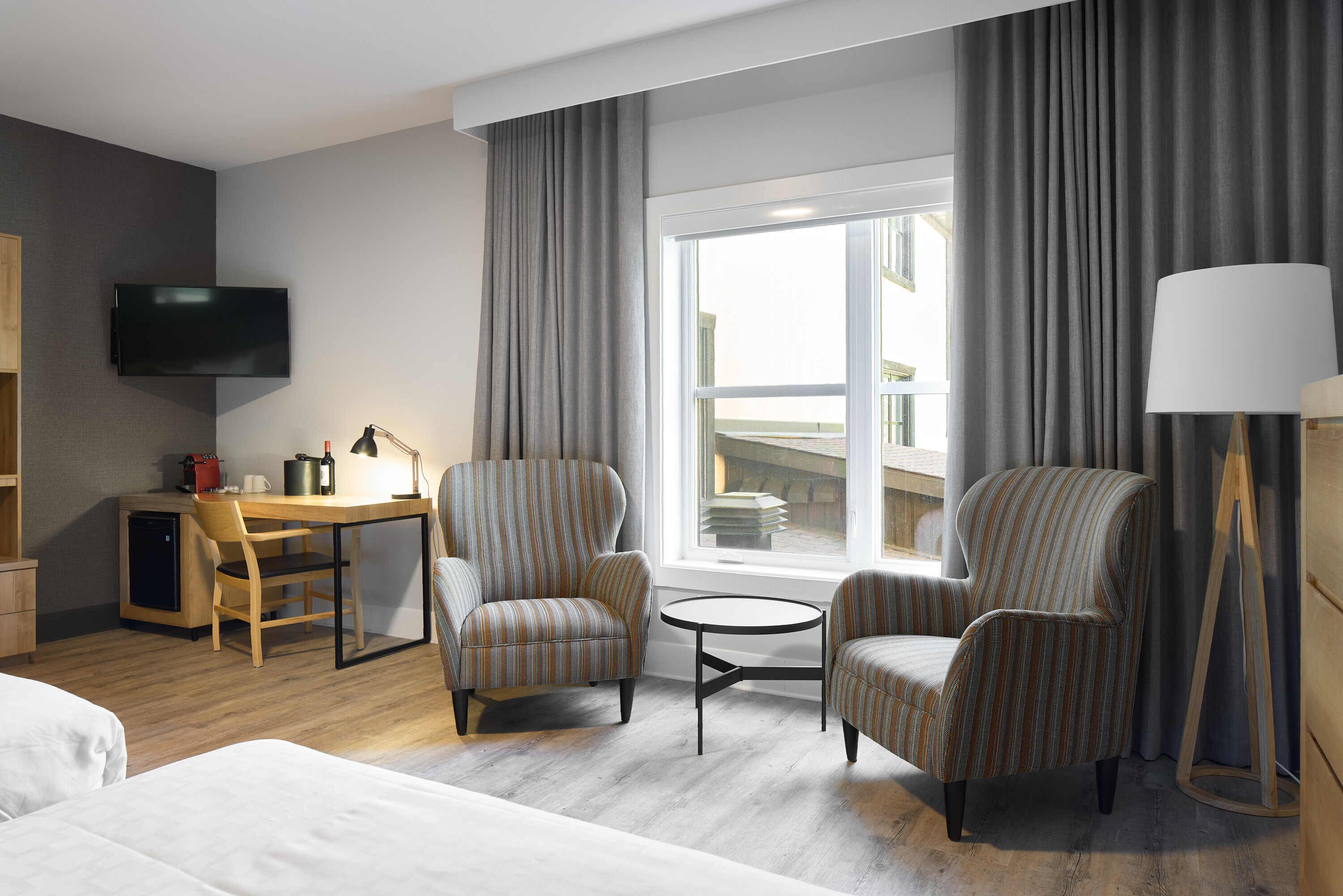 Habitación de hotel con cama, escritorio, dos sillones a rayas, una mesita, televisor y ventana con cortinas.