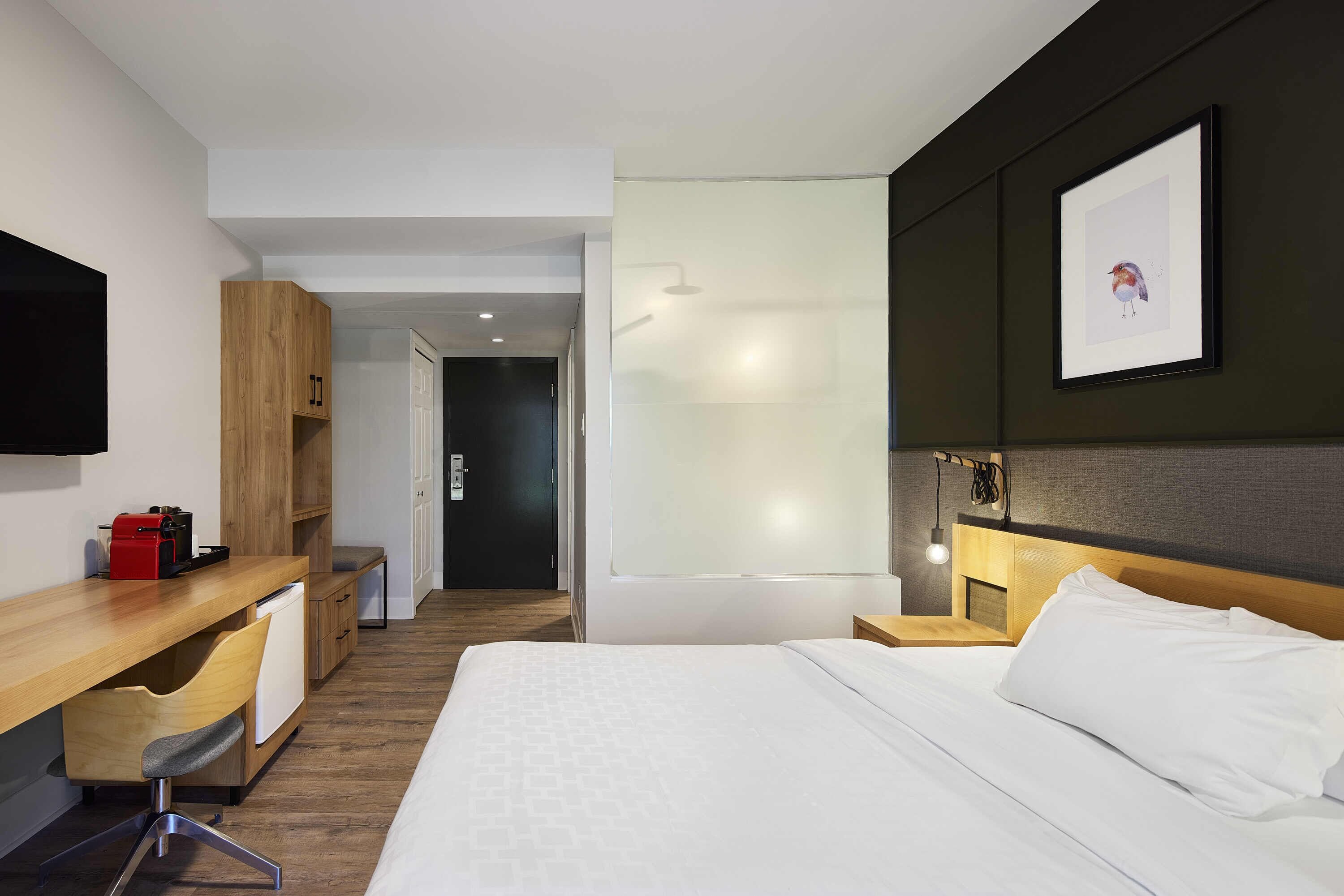 Una habitación de hotel con una cama, un escritorio con una silla, una mesita pequeña y un televisor montado en la pared.