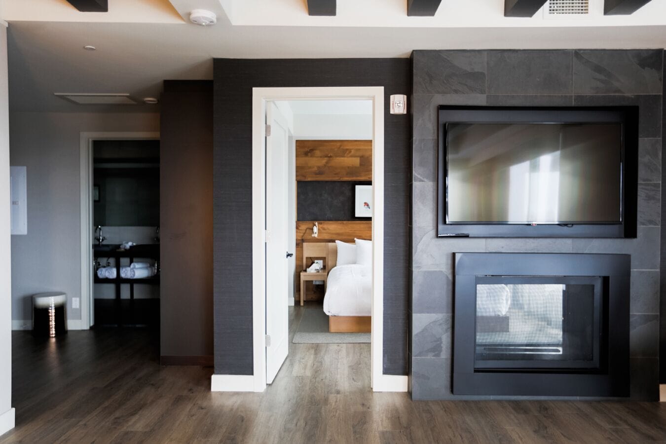 A modern hotel room with a dark wood floor, a large flat-screen TV mounted above a fireplace, and a wooden headboard in the background.