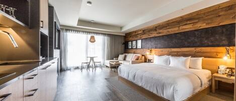 A modern hotel room with a large bed, a wooden accent wall, a dining area with a table and chairs, and a kitchen area with a built-in oven.