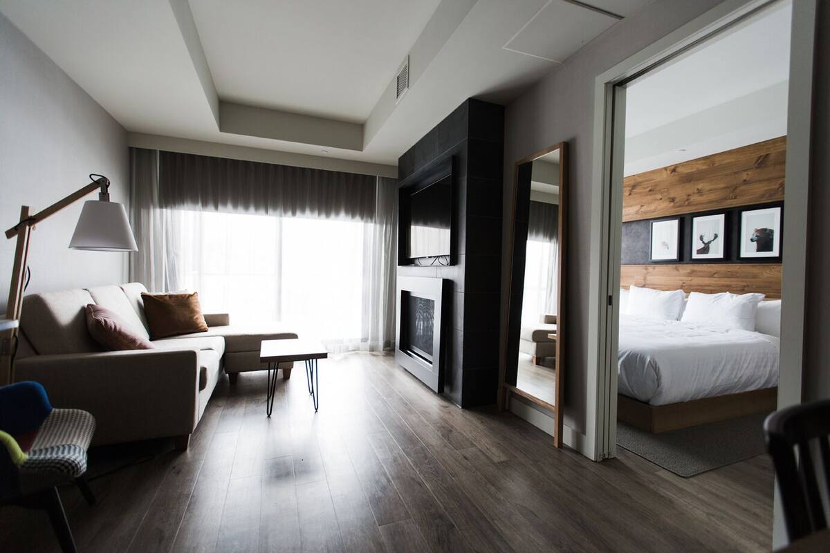 A modern hotel room with a sofa, a small table, a lamp, a bed, and a wooden accent wall.