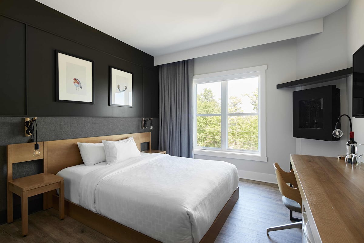 A modern hotel room with a large bed, a wooden desk, and a window with a view of greenery.