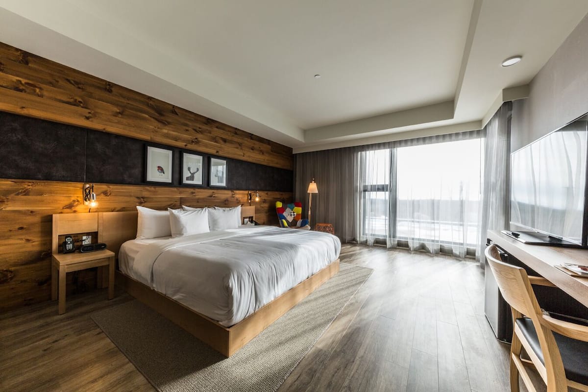 A modern hotel room with a large bed, wooden headboard, bedside tables with lamps, a desk with a chair, and a large window with curtains.