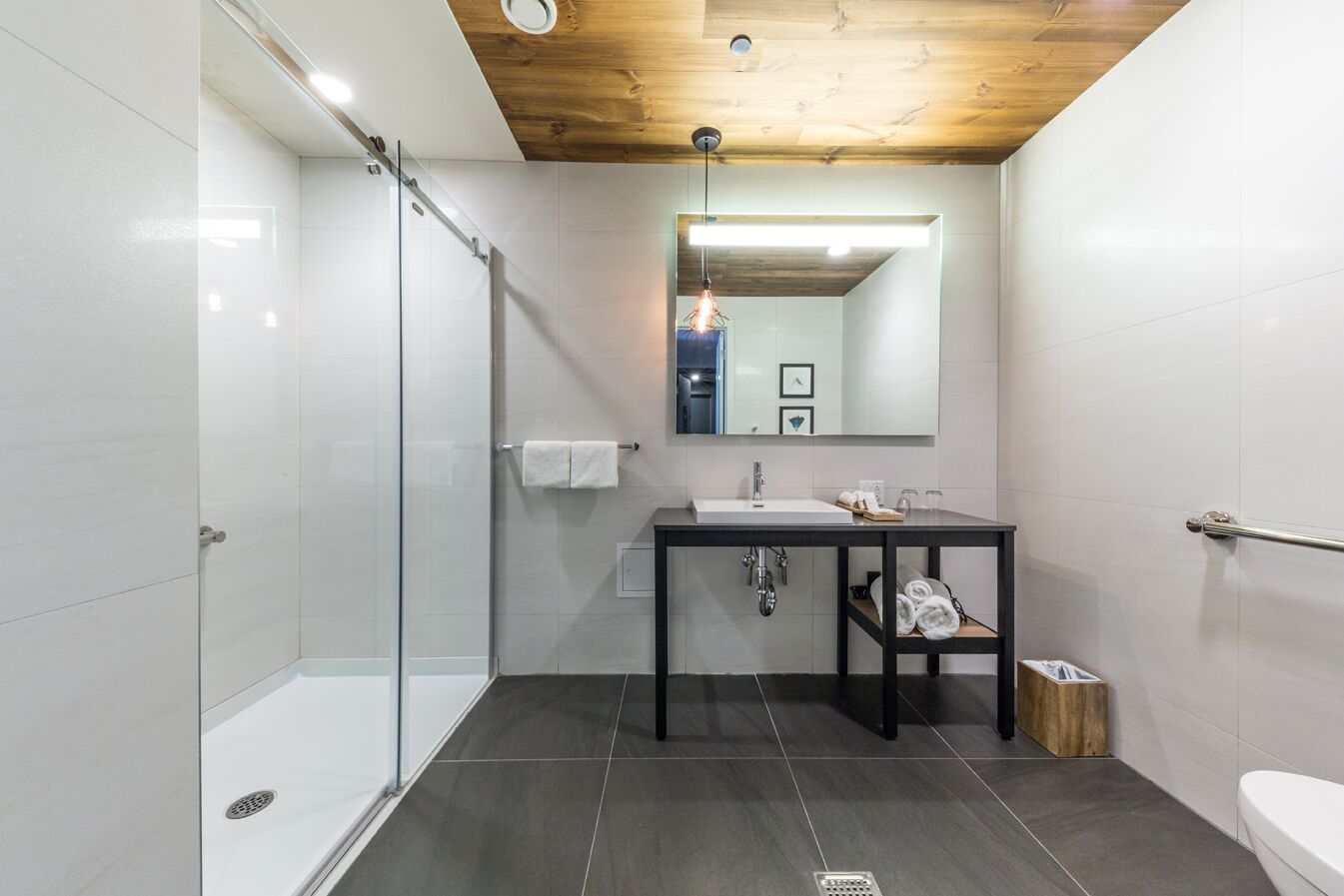 Un baño moderno con cabina de ducha de vidrio, suelo de baldosas oscuras, un lavamanos blanco con espejo sobre él y techo de madera.